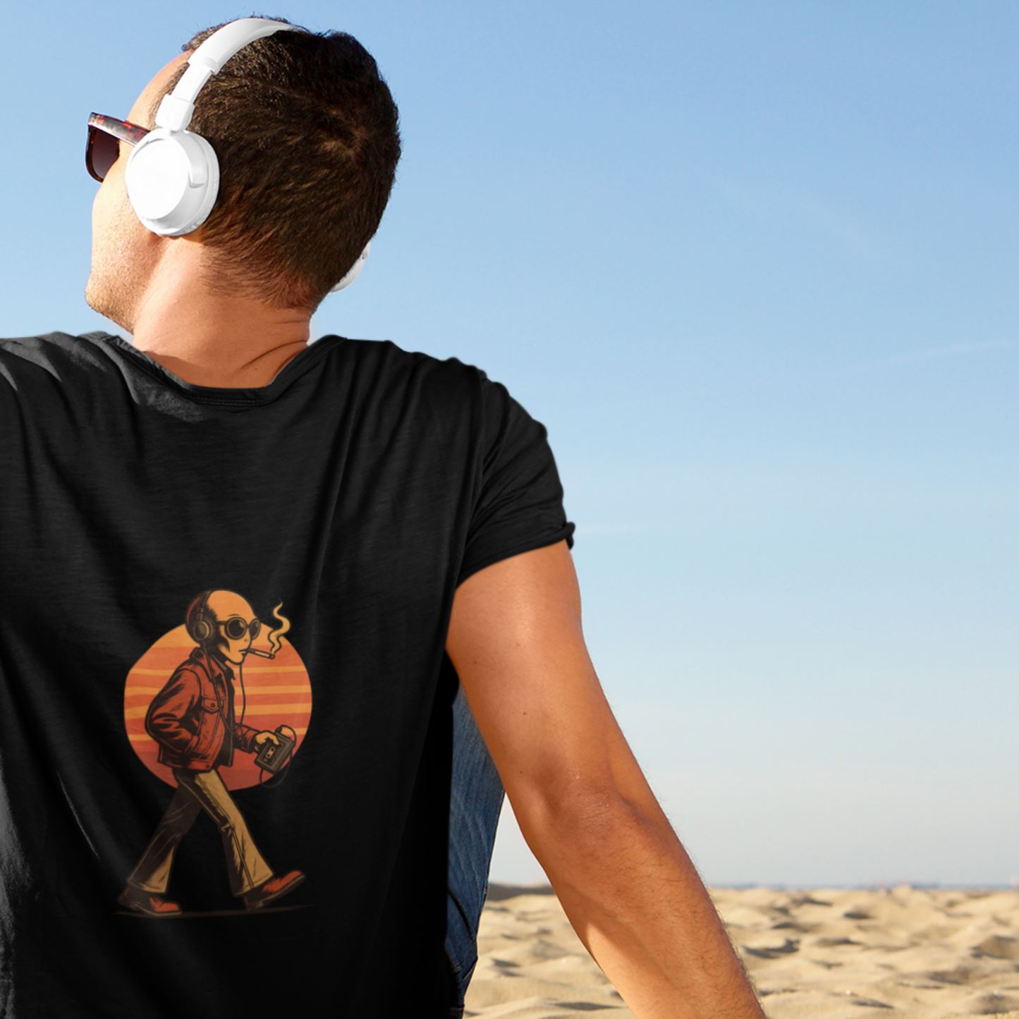 Man sitting on sand wearing black t-shirt with retro alien graphic, headphones and sunset design on back
