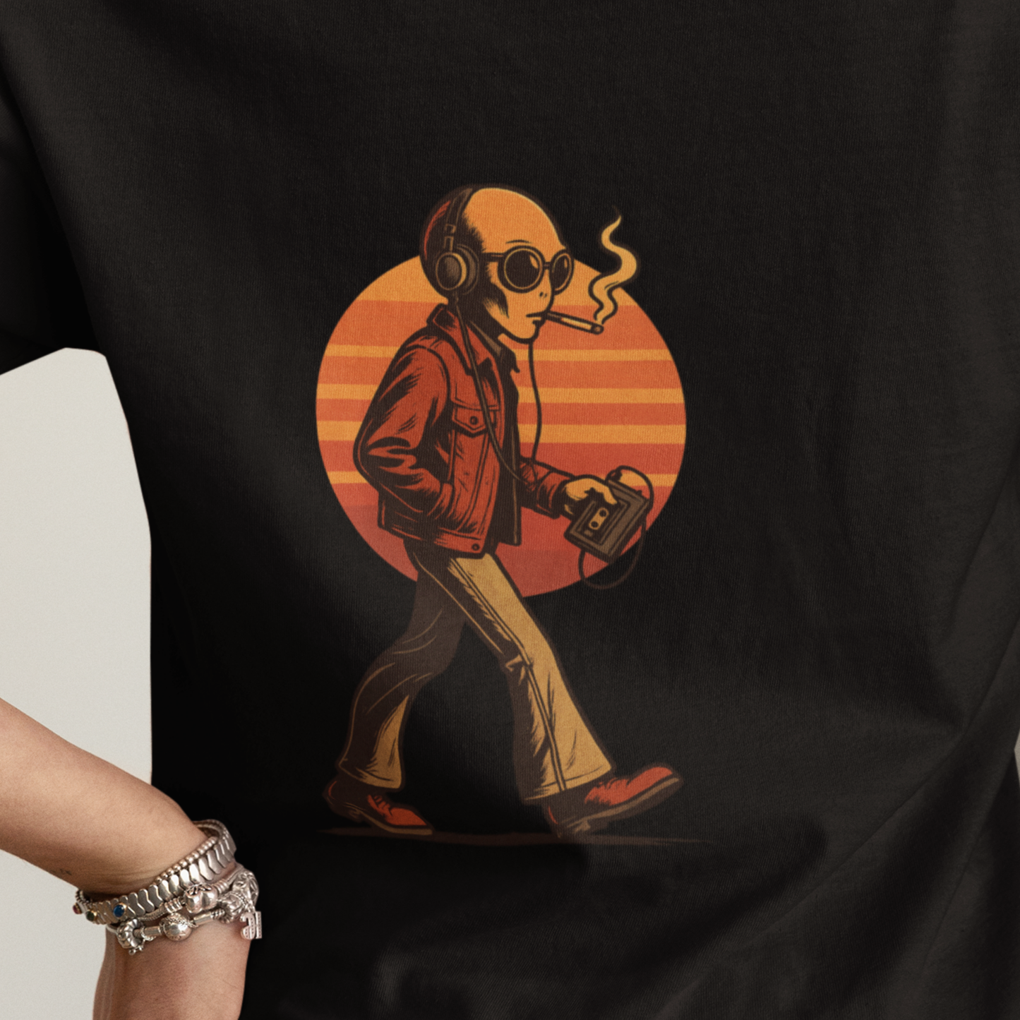 Close-up of retro smoking alien graphic with Walkman on black cotton t-shirt, orange sunset background behind design.