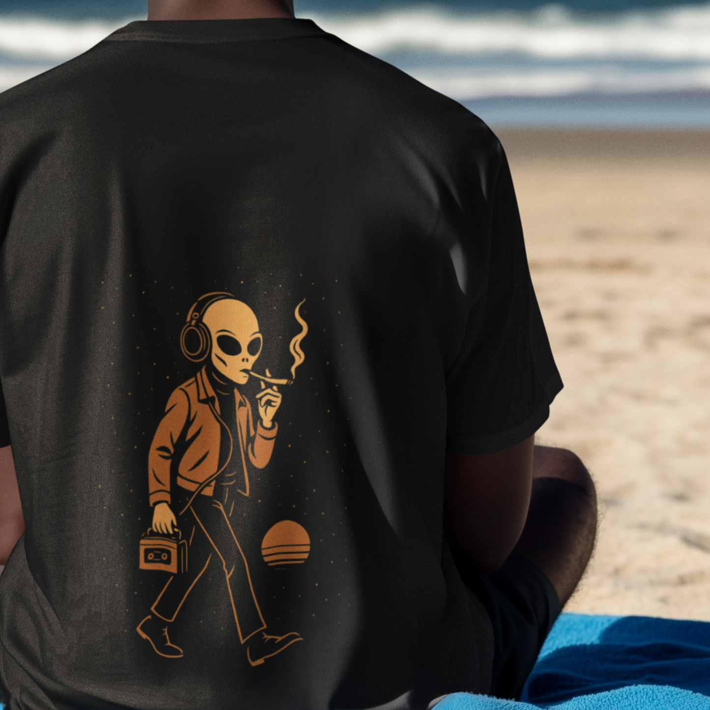 Close-up of a back-printed black t-shirt featuring a retro alien design, worn by a model seated on a beach with a calm ocean backdrop.