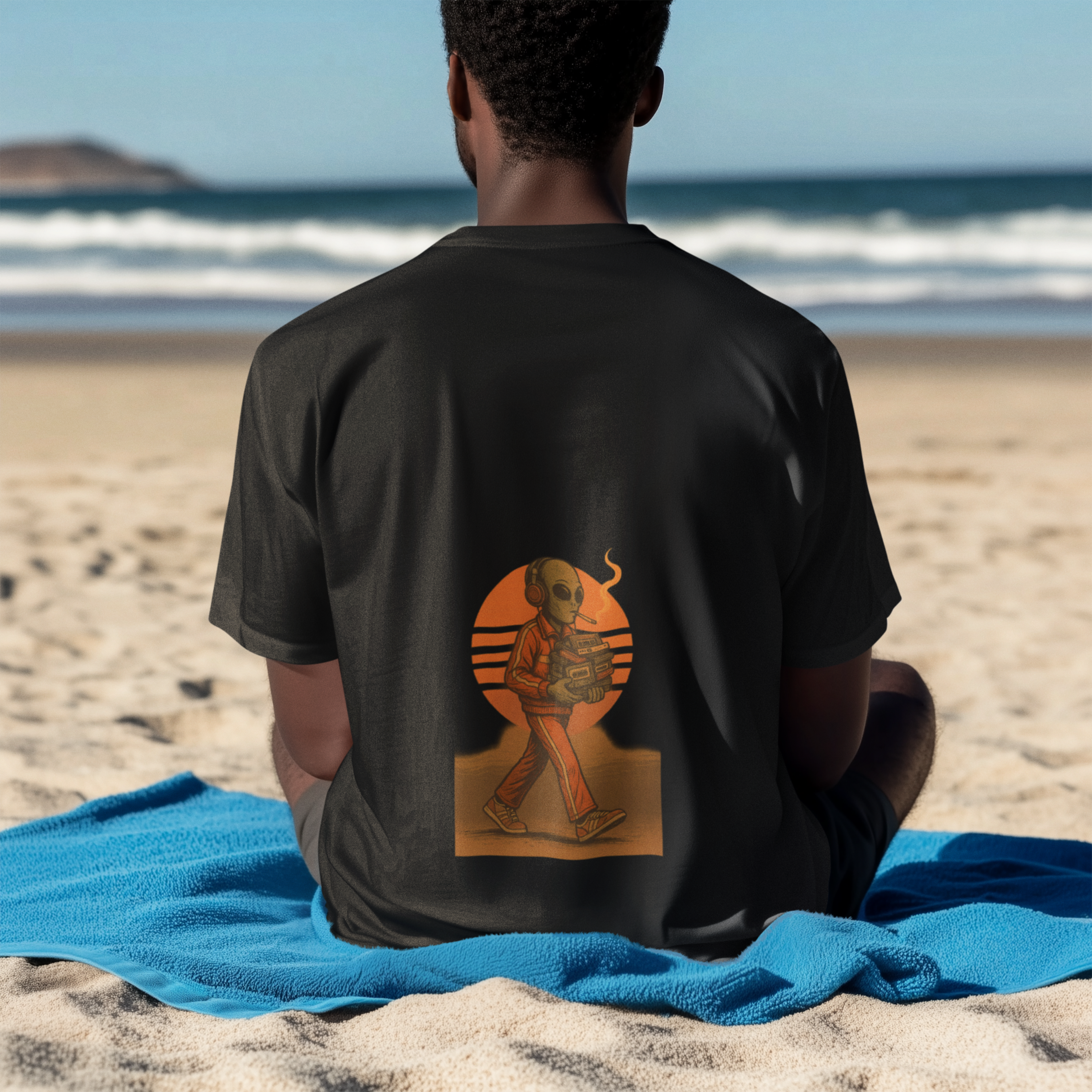 Man sitting on beach wearing black t-shirt with back print of a smoking alien holding a cassette player against an orange retro sun.