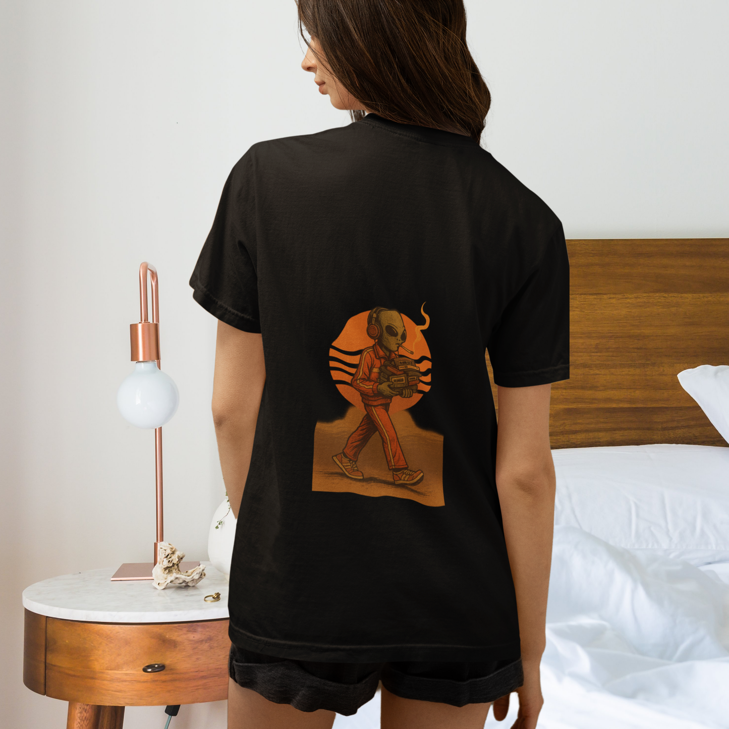 Woman wearing black t-shirt with back print of a smoking alien and sunset design, standing beside a modern bedside lamp and wooden headboard.