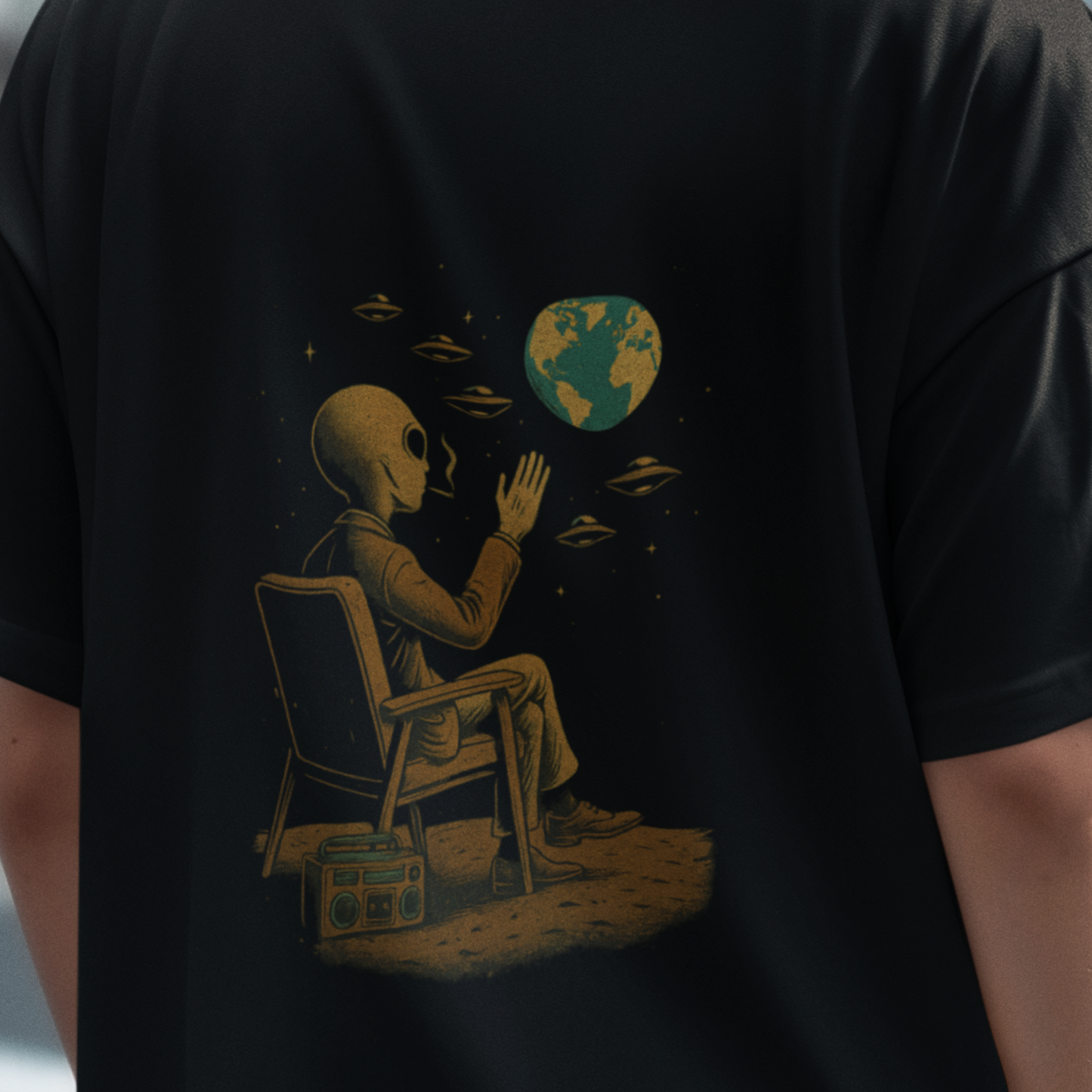 Close-up of a black oversized t-shirt showing an alien sitting on a chair, watching Earth with flying saucers in the sky. Design printed on the back of a Comfort Colors 1717 tee.