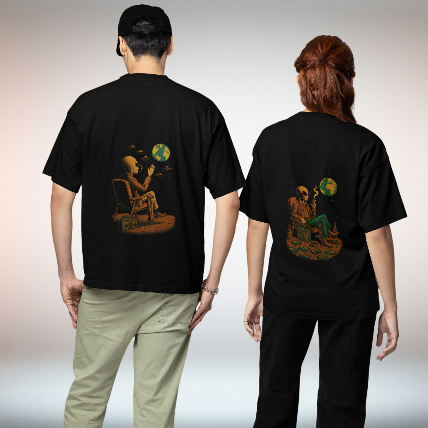 Two models wearing black oversized t-shirts with back prints of an alien sitting on a chair, watching Earth. Unisex Comfort Colors 1717 fit shown from behind.