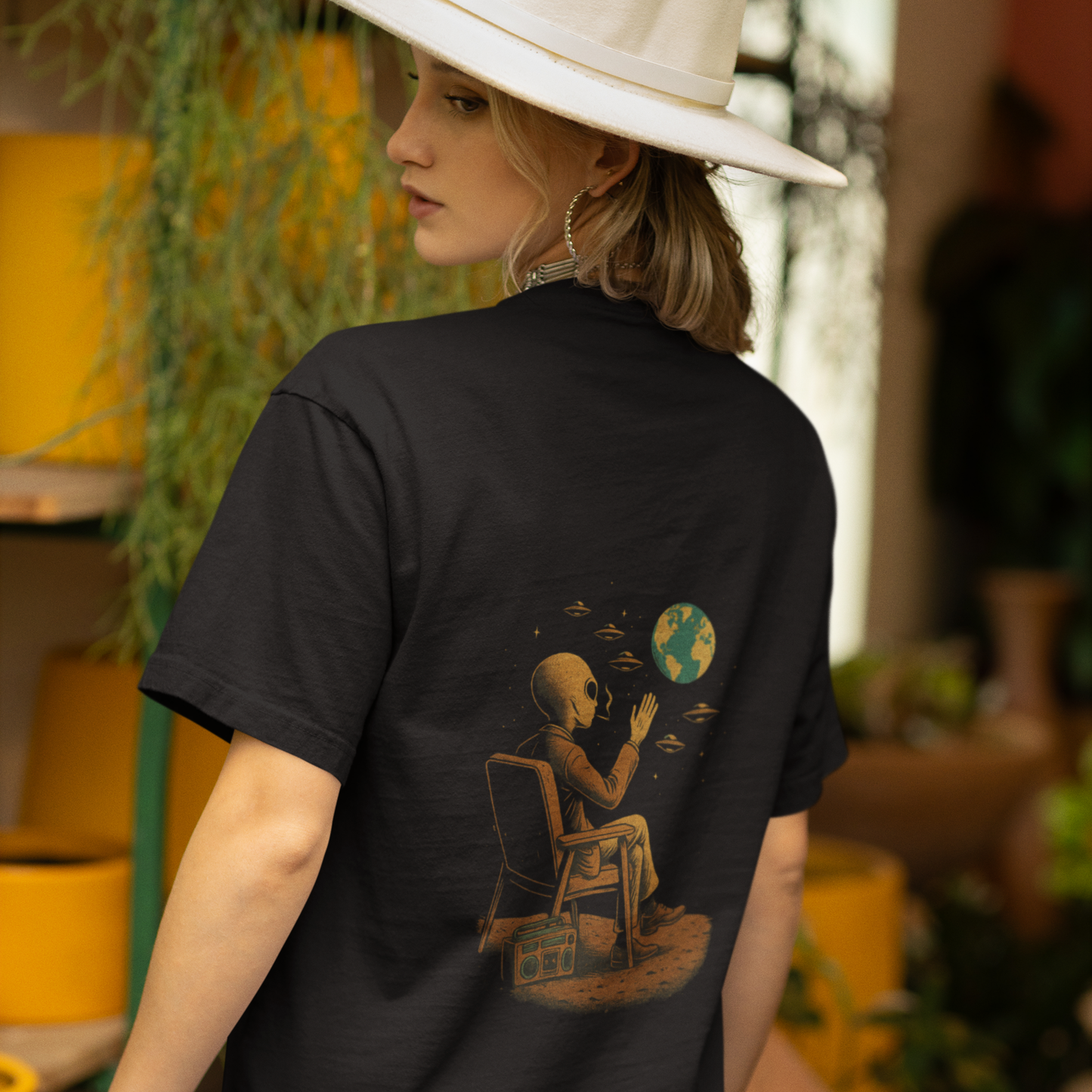 Back view of a woman wearing a black oversized t-shirt with an alien and Earth back print design, standing in a plant-filled indoor setting. Printed on Comfort Colors 1717 tee.