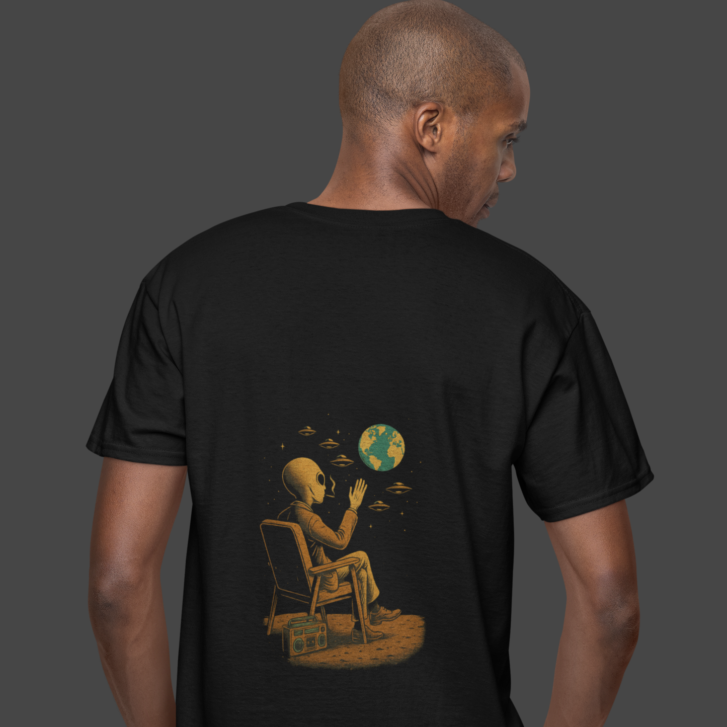 Back view of a man wearing a black oversized t-shirt with an alien and Earth graphic print on the back. T-shirt shown on neutral studio background. Printed on Comfort Colors 1717.