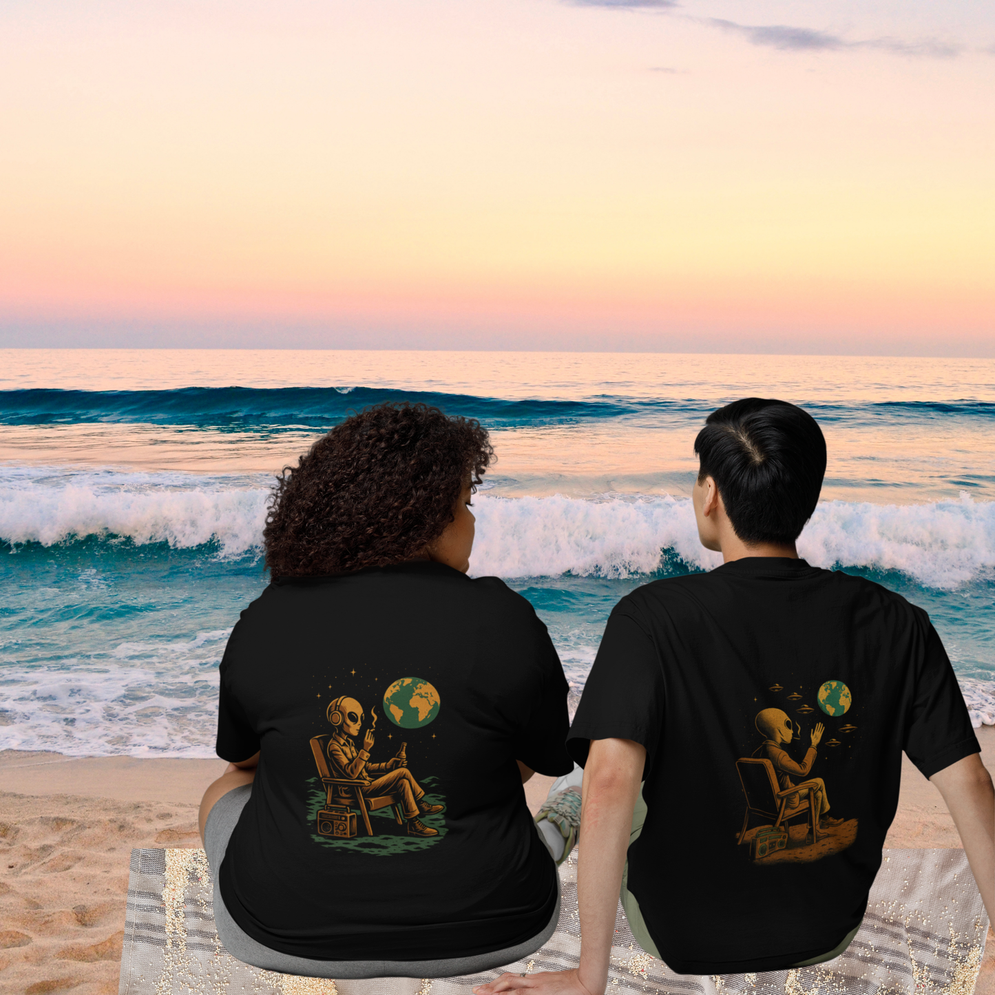 Two people sitting on the beach wearing black oversized t-shirts with back prints of an alien watching Earth. Sunset ocean view highlights the relaxed, cosmic vibe.