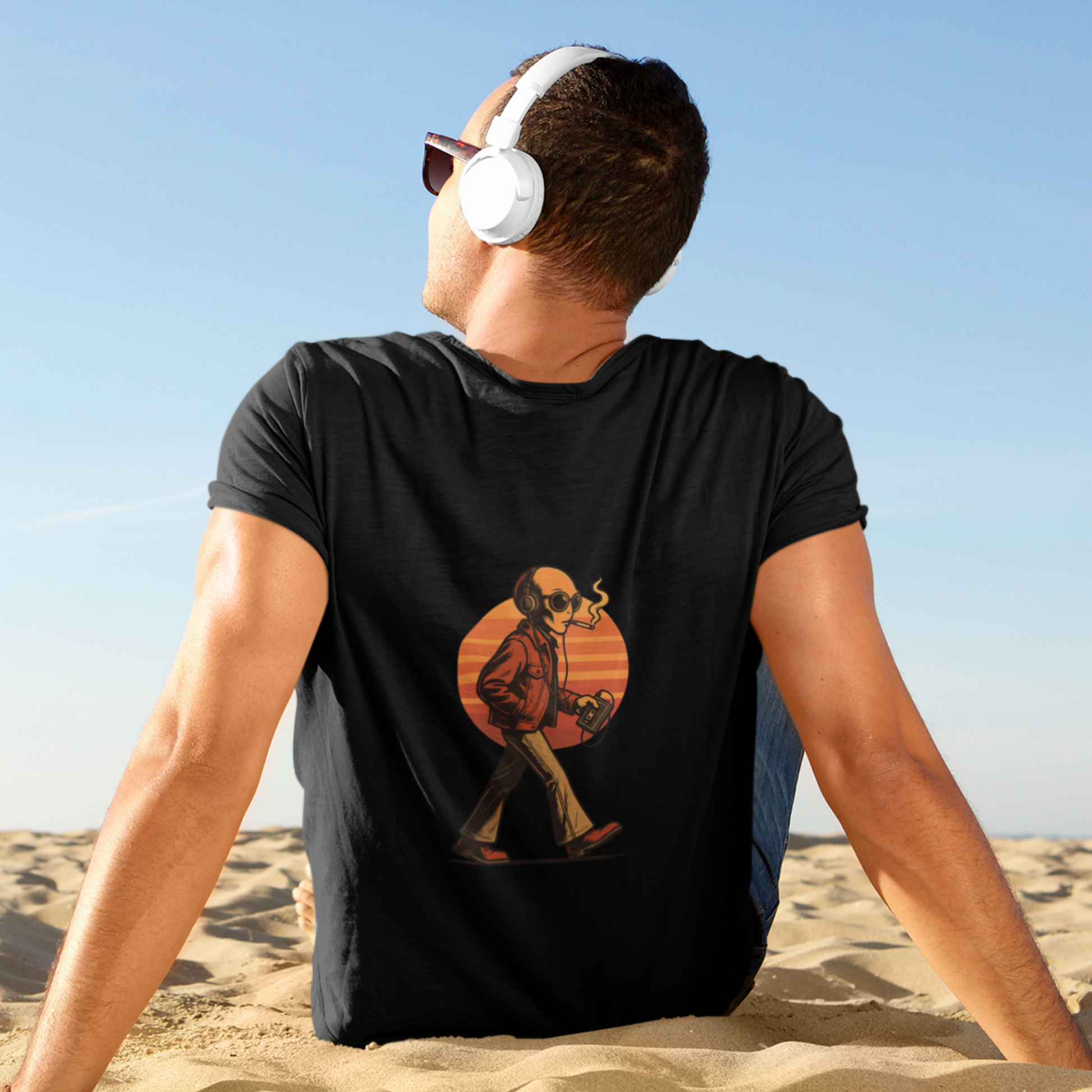 Man wearing black t-shirt with retro alien back print, sitting on the beach with headphones on and facing the sky