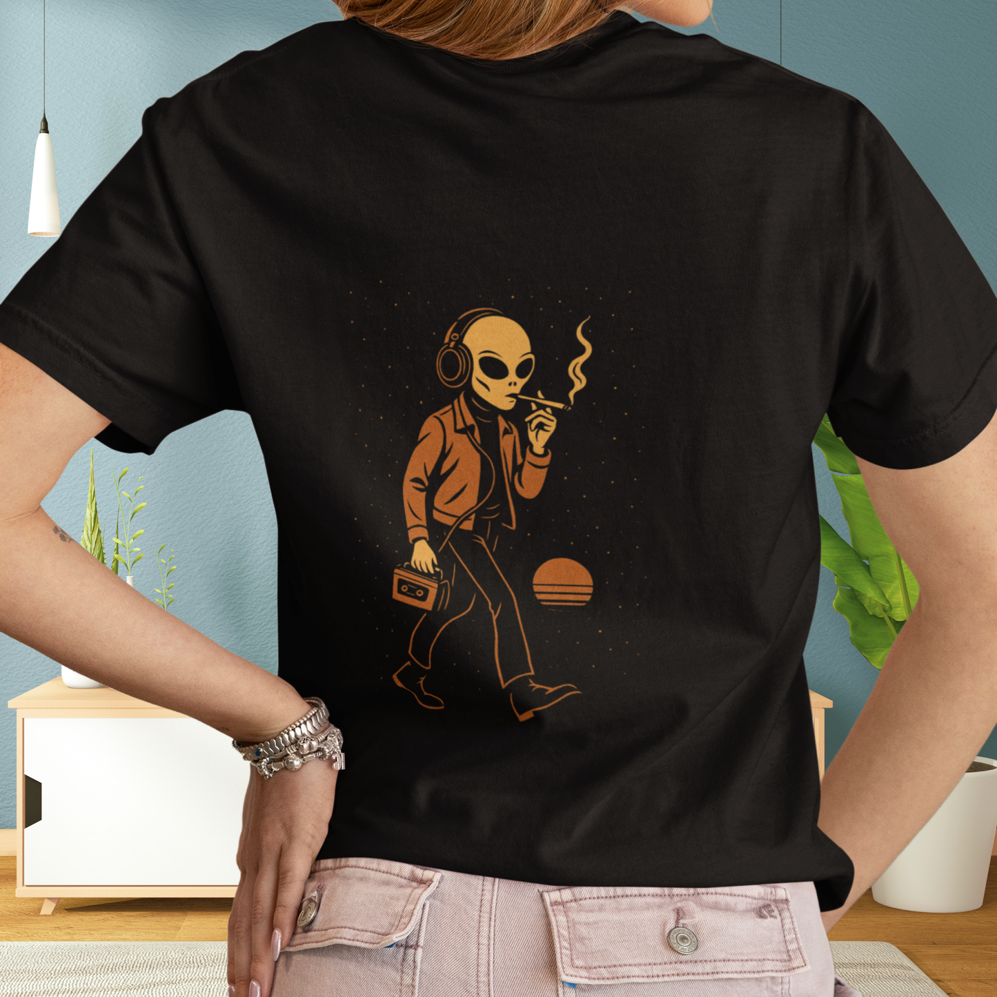 Woman wearing a black t-shirt featuring a back print of a smoking alien with headphones and cassette player, standing in a cozy modern interior.