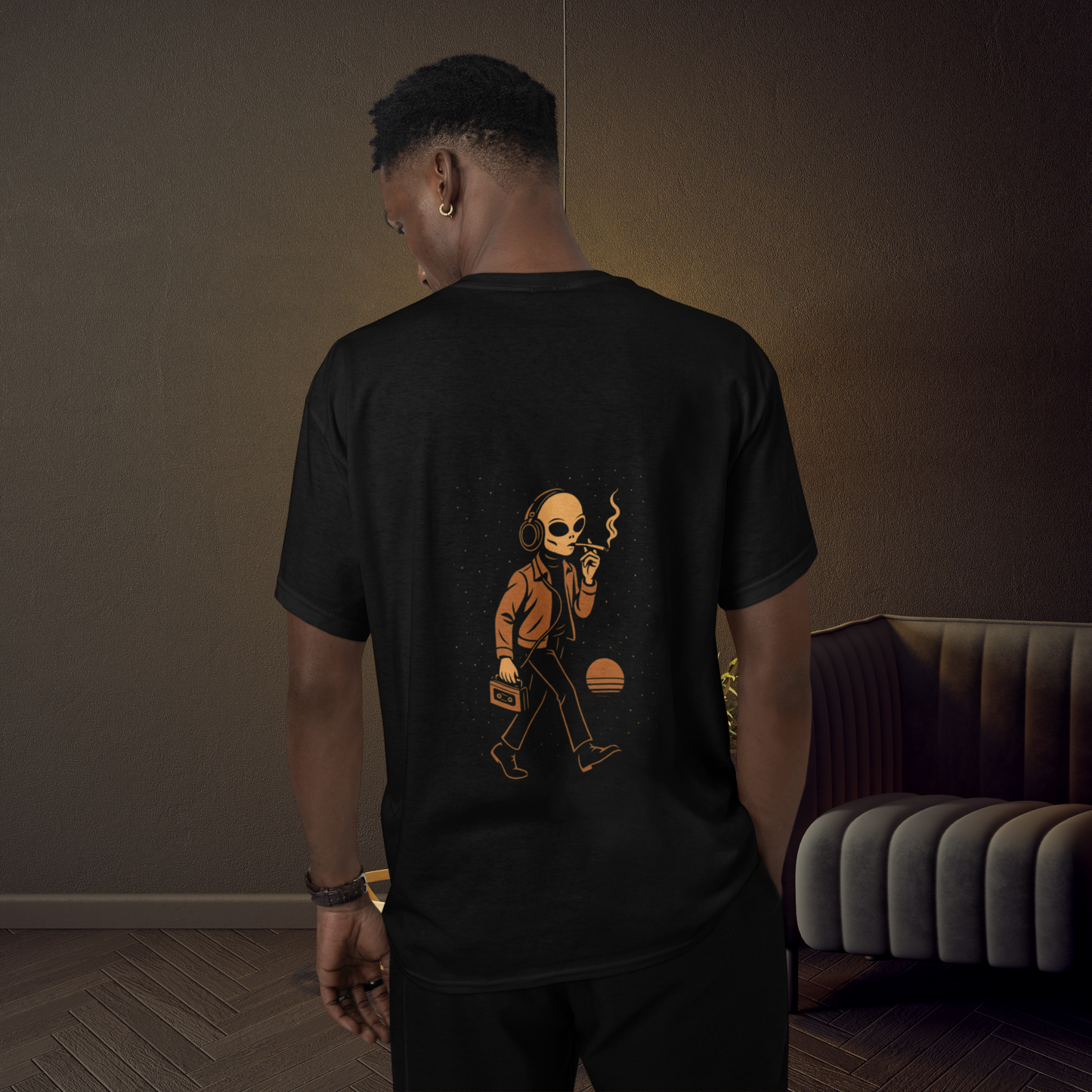 Black unisex t-shirt featuring a back-printed graphic of a smoking alien with headphones and a cassette player, standing indoors with moody ambient lighting.