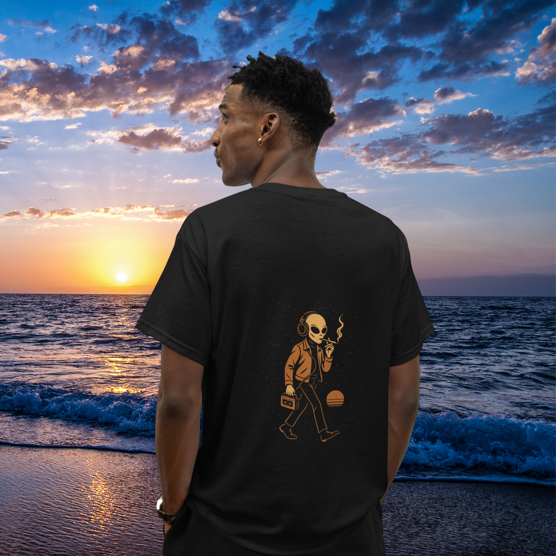 Model standing by the ocean during sunset, wearing a black t-shirt with a back print of a smoking alien in headphones holding a cassette player.