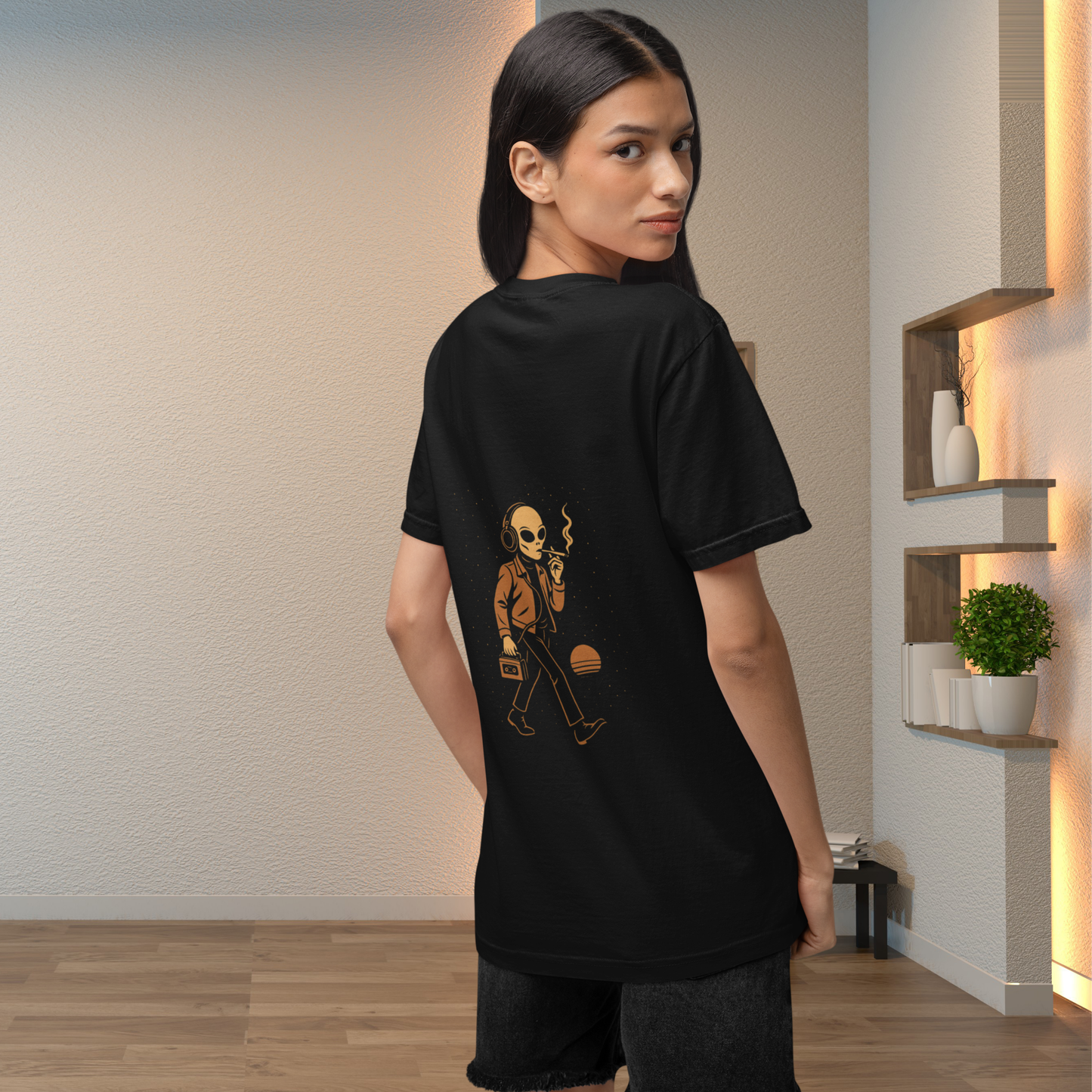 Female model wearing a black unisex t-shirt with a retro alien back print design, photographed indoors in warm modern lighting.