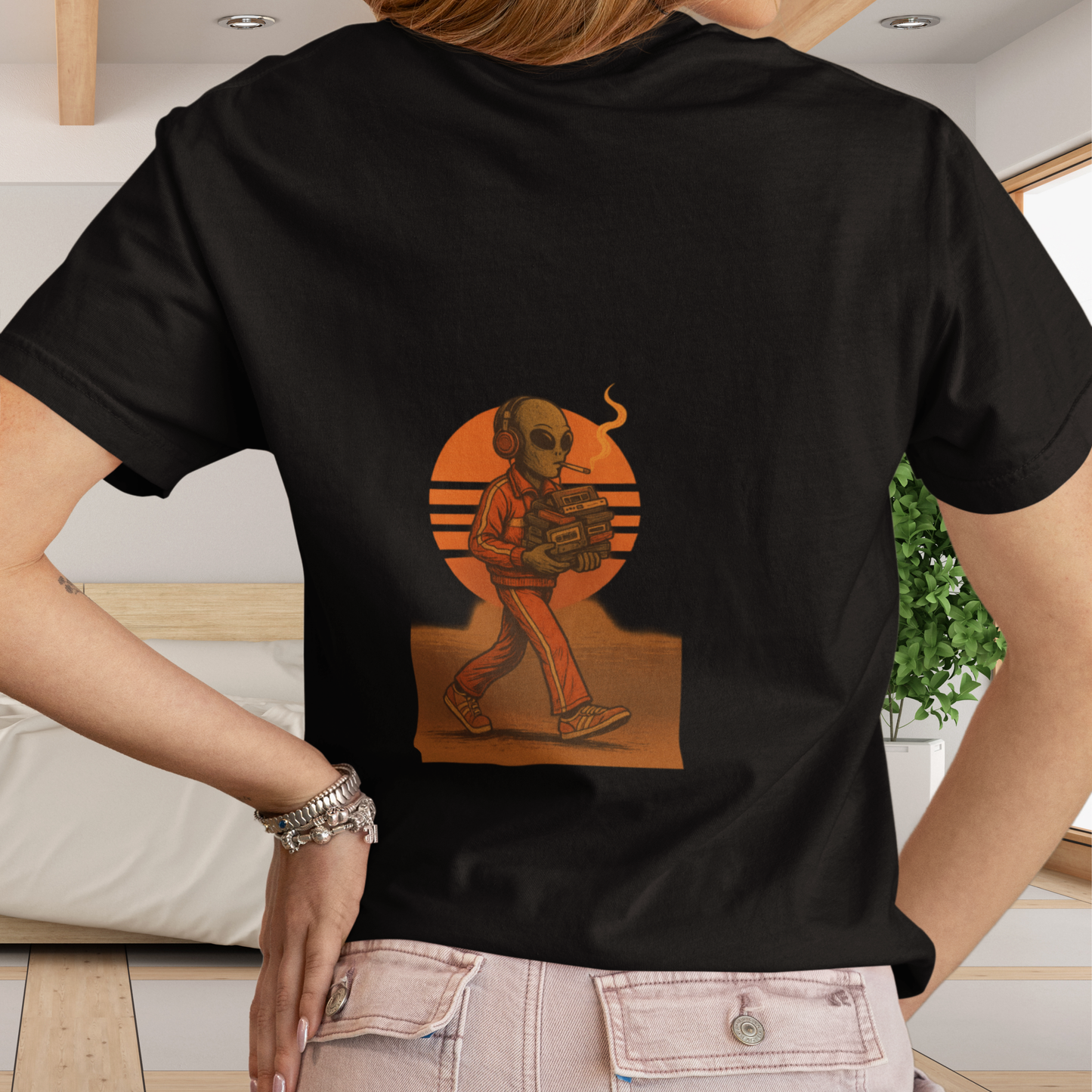 Woman wearing black t-shirt with back graphic of a smoking alien holding a cassette player, walking in front of a retro orange sun design.