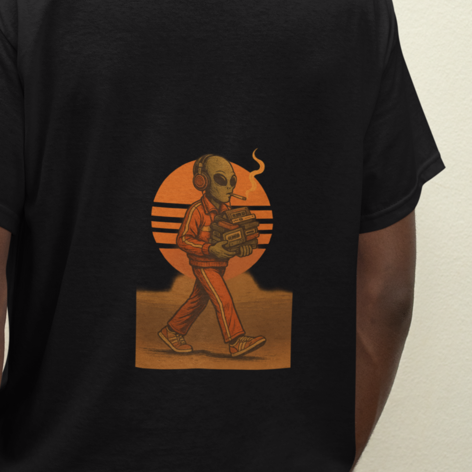 Close-up of black t-shirt featuring a back print of a smoking alien in headphones, holding a boombox, walking in front of an orange retro sunset.