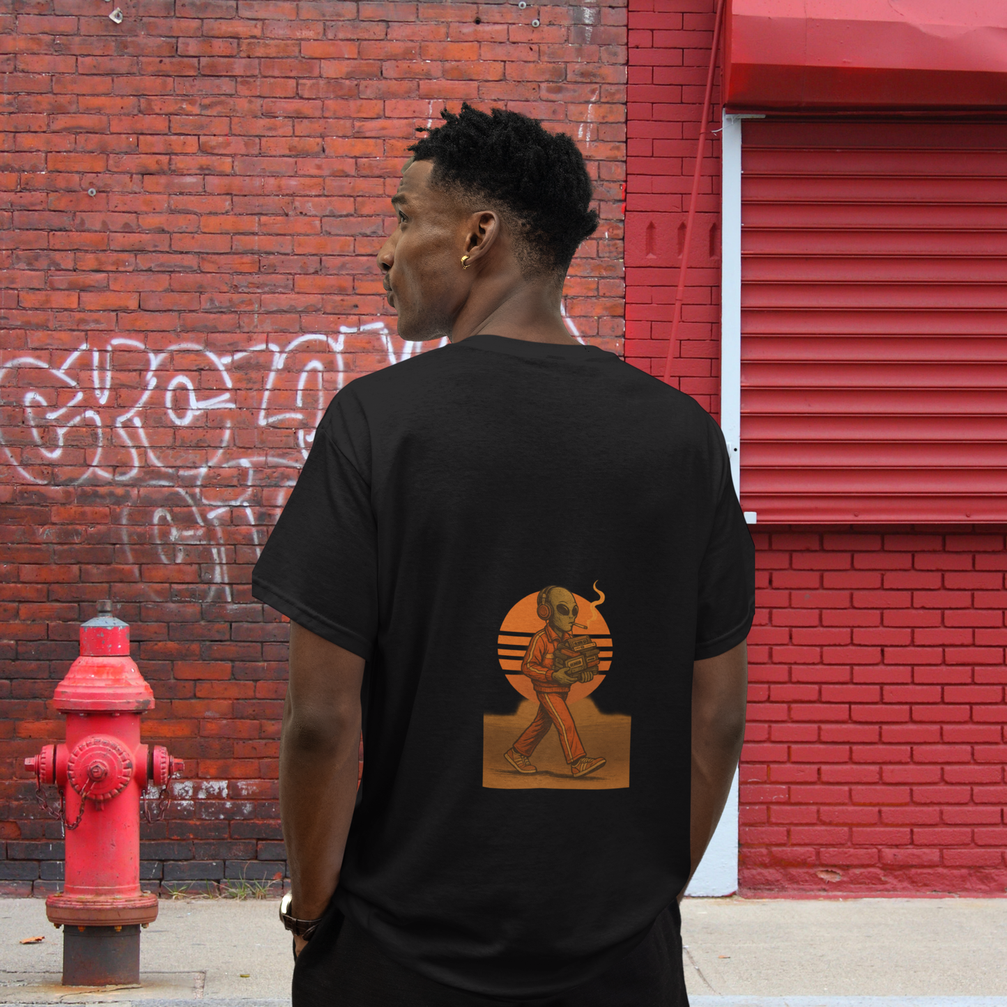 Man wearing black t-shirt with back print of a retro-style smoking alien in front of an orange sunset, standing against urban red brick wall.